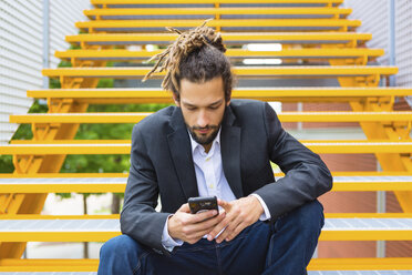 Junger Geschäftsmann mit Dreadlocks sitzt auf einer Treppe und benutzt ein Smartphone - MGIF00015