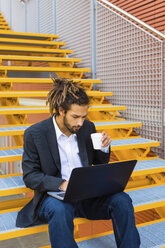 Junger Geschäftsmann mit Dreadlocks sitzt auf einer Treppe und benutzt einen Laptop - MGIF00016