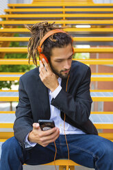 Junger Geschäftsmann mit Dreadlocks sitzt auf einer Treppe und hört Musik mit Kopfhörern und Handy - MGIF00017