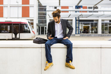 Junger Geschäftsmann mit Dreadlocks benutzt sein Smartphone beim Warten am Bahnhof - MGIF00021