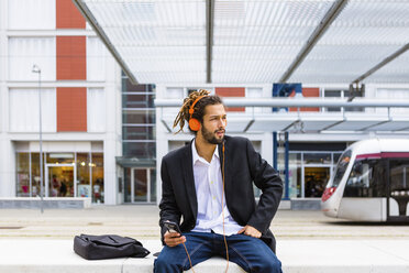 Junger Geschäftsmann mit Dreadlocks hört Musik mit Kopfhörern und Handy am Bahnhof - MGIF00024