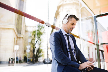 Businessman with smartphone and headphones waiting at the bus stop - HAPF01746