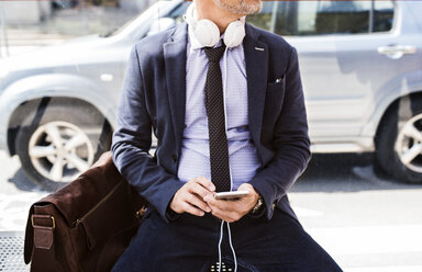 Businessman with smartphone and headphones waiting at the bus stop - HAPF01749