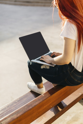 Rothaarige Frau sitzt auf einer Bank und benutzt einen Laptop, lizenzfreies Stockfoto