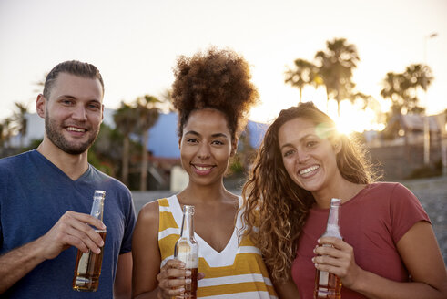 Gruppenbild von drei Freunden mit Bierflaschen am Strand bei Sonnenuntergang - PACF00038