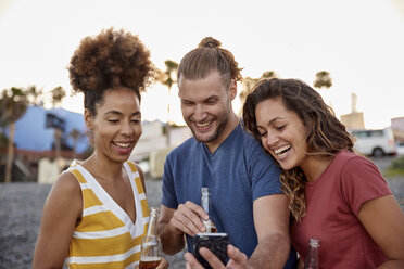 Three laughing friends with beer bottles looking at smartphone on the beach - PACF00048