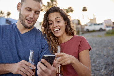 Pärchen mit Bierflaschen schaut auf Handy am Strand - PACF00049