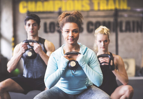 Selbstbewusste junge Frau mit Trainingspartnern, die eine Kettlebell im Fitnessstudio heben - HAPF01832