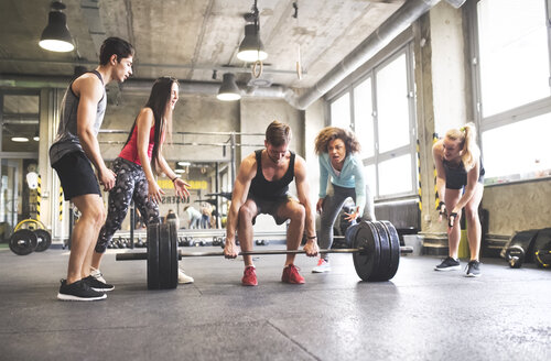 Gruppe junger fitter Leute, die einen Mann beim Gewichtheben im Fitnessstudio anfeuern - HAPF01838