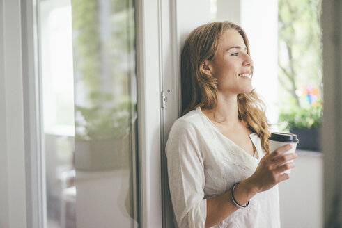 Lächelnde junge Frau mit Kaffee zum Mitnehmen am Fenster - KNSF01631