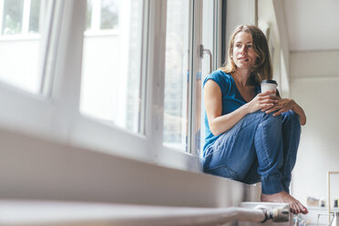 Lächelnde junge Frau mit Kaffee zum Mitnehmen, die am Fenster sitzt - KNSF01641