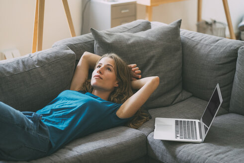 Junge Frau mit Laptop zu Hause auf der Couch liegend - KNSF01650