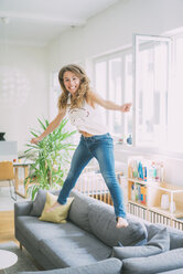 Excited young woman jumping on couch at home - KNSF01652