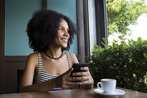 Junge Frau mit Smartphone sitzt in einem Café und schaut aus dem Fenster - ABZF02147