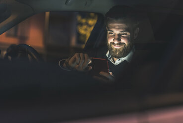 Lächelnder Geschäftsmann, der nachts im Auto ein Mobiltelefon benutzt - UUF10882