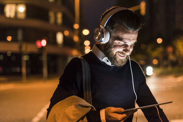 Lächelnder junger Mann mit Tablet und Kopfhörern auf einer nächtlichen Straße in der Stadt - UUF10889