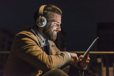 Lächelnder junger Mann mit Tablet und Kopfhörern in der Stadt bei Nacht - UUF10893