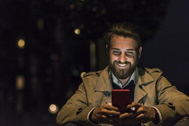 Lächelnder junger Mann mit Mobiltelefon in der Stadt bei Nacht - UUF10896