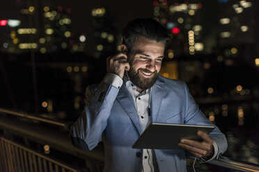 Lächelnder junger Mann mit Tablet und Kopfhörer auf städtischer Brücke bei Nacht - UUF10904