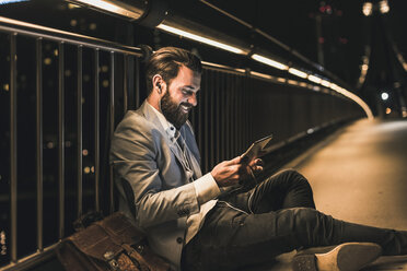 Smiling young man with tablet and earphone sitting on bridge at night - UUF10906