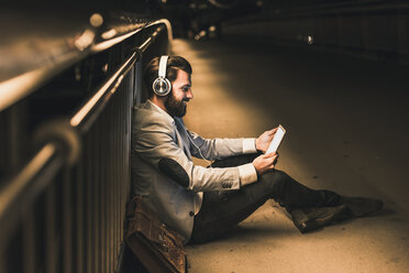 Smiling young man with tablet and headphones sitting on bridge at night - UUF10907