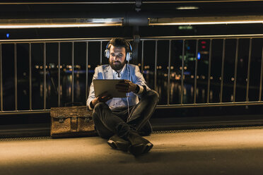 Lächelnder junger Mann mit Tablet und Kopfhörern, der nachts auf einer Brücke sitzt - UUF10908