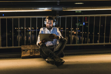 Smiling young man with tablet and headphones sitting on bridge at night - UUF10908