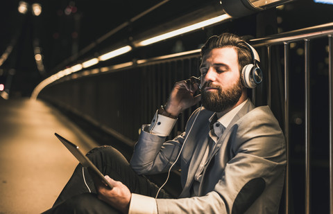 Junger Mann mit Tablet und Kopfhörern sitzt nachts auf einer Brücke, lizenzfreies Stockfoto