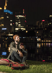 Smiling young man with guitar and cell phone sitting at urban riverside at night - UUF10910