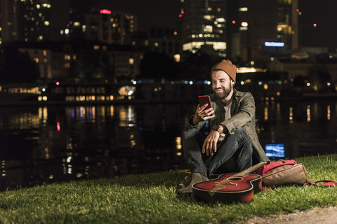 Lächelnder junger Mann mit Gitarre und Mobiltelefon am nächtlichen Flussufer, lizenzfreies Stockfoto