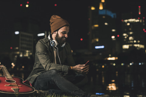 Lächelnder junger Mann mit Gitarre, Handy und Kopfhörern, der am nächtlichen Flussufer sitzt, lizenzfreies Stockfoto