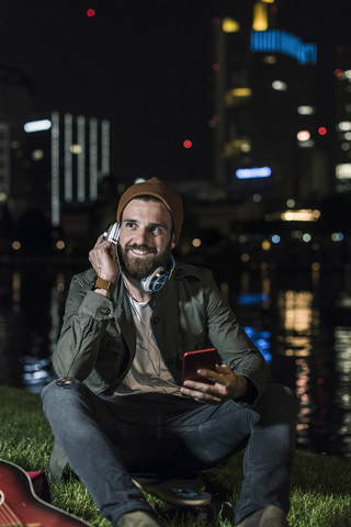 Lächelnder junger Mann mit Gitarre, Handy und Kopfhörer am nächtlichen Flussufer sitzend, lizenzfreies Stockfoto