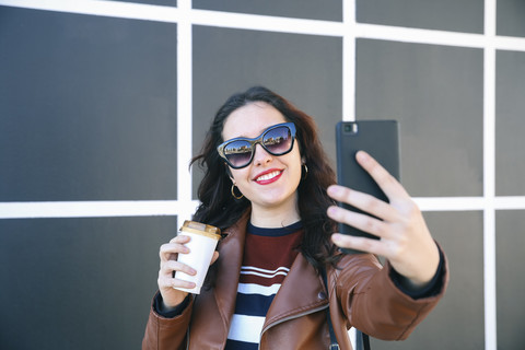 Porträt einer lächelnden jungen Frau mit Kaffee zum Mitnehmen, die ein Selfie mit ihrem Smartphone macht, lizenzfreies Stockfoto
