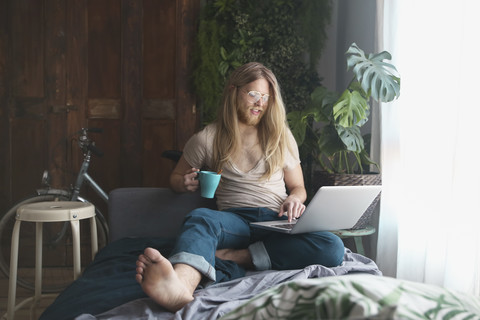 Bärtiger junger Mann mit langen Haaren, der einen Laptop benutzt und zu Hause Kaffee trinkt, lizenzfreies Stockfoto