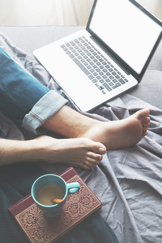 Mann auf Schlafsofa liegend mit Laptop und Tasse Kaffee, Teilansicht, lizenzfreies Stockfoto