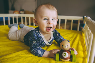 Portrait of baby boy with wooden toy lying in his crib - MFF03676