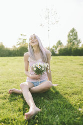 Lachende Frau, die auf einer Wiese sitzt und einen Strauß Gänseblümchen hält - GIOF02829