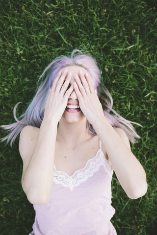 Lachende Frau im Gras liegend, die ihre Augen mit den Händen bedeckt, lizenzfreies Stockfoto