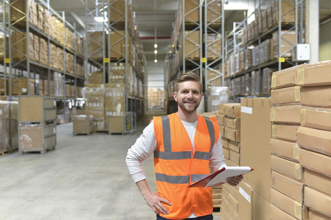 Porträt eines lächelnden Mannes in einer Fabrikhalle, der eine Sicherheitsweste trägt und ein Klemmbrett hält, lizenzfreies Stockfoto