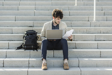 Nachdenklicher Mann mit Laptop, der auf einer Treppe sitzt und Dokumente überprüft - GIOF02886