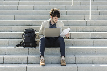 Pensive man with laptop sitting on steps checking documents - GIOF02886