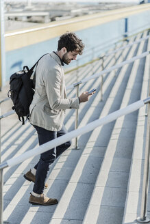 Lächelnder Geschäftsmann, der die Treppe hinaufgeht und auf sein Mobiltelefon schaut - GIOF02888