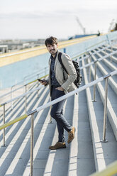 Portrait of businessman with backpack and cell phone walking downstairs - GIOF02889