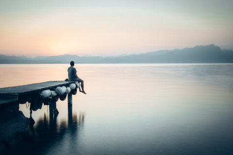 Italien, Lazise, Mann sitzt auf Steg und schaut auf den Gardasee, lizenzfreies Stockfoto