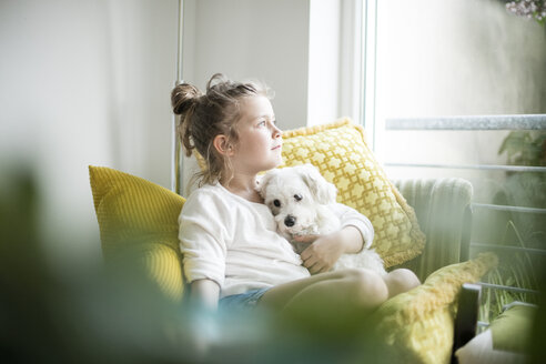 Kleines Mädchen sitzt auf einem Sessel zu Hause und kuschelt mit ihrem Hund - MOEF00053