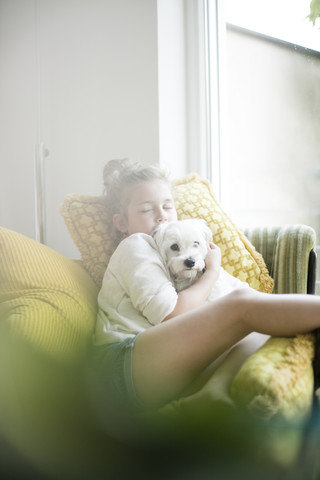 Kleines Mädchen sitzt auf einem Sessel zu Hause und kuschelt mit ihrem Hund, lizenzfreies Stockfoto