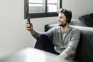 Smiling young man with cell phone and headphones at home - GIOF02896