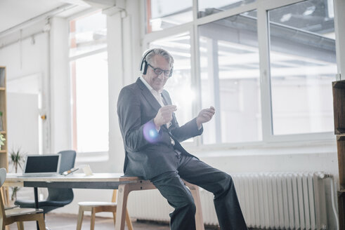 Happy senior businessman listening to music with headphones in office - GUSF00040