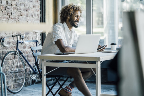 Lächelnder junger Mann am Schreibtisch mit Laptop, lizenzfreies Stockfoto