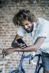 Glücklicher junger Mann mit Fahrrad, der sein Mobiltelefon überprüft - KNSF01740
